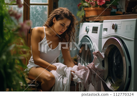 Young woman in a casual outfit sorting clothes for washing in a cozy laundry room 112770324
