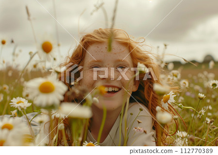 Happy young girl with red hair enjoys a playful moment among wild daisies 112770389