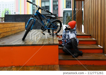 A Caucasian boy dressed in a winter jacket walks with a bicycle around the city 112771261