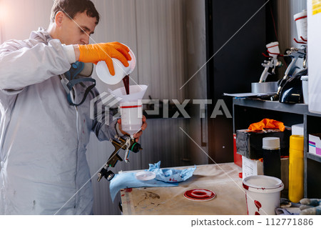 Male car painter in protective clothes pouring mixed paint from a can into a plastic mixing bucket at car service station. 112771886