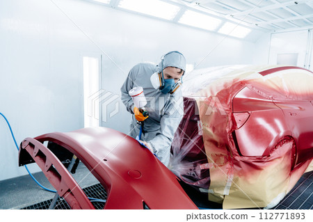Male car painter in protective clothes and mask painting automobile bumper with red paint and varnish in chamber workshop. 112771893
