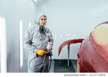 Automobile repairman painter hand in protective glove with airbrush pulverize painting car body in paint chamber. Automobile repairman painter hand in protective glove with airbrush pulverize painting car body in paint chamber. 112771903
