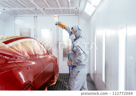 Automotive paint services, quality auto body shop concept. Real shot of man of professional car painter with protective clothing standing next to car in auto paint room and with gun 112772084