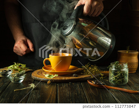 A man adds boiling water to a mug to make healthy tea on the kitchen table. Traditional medicine concept or preparation of medicinal tea with medicinal dry herbs 112773847