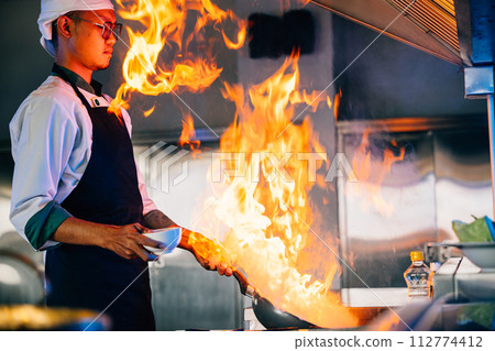 In professional kitchen chef hands handle flaming wok. Closeup of cooking expertise flames at work. Skillful chef burning food working in a modern kitchen setting. cook food with fire 112774412