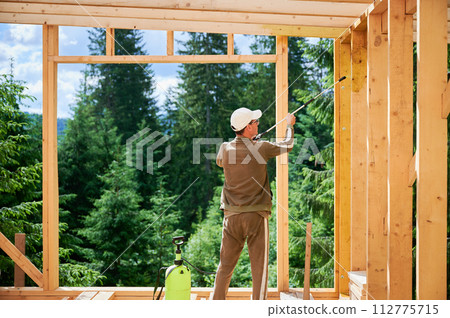 Laborer constructing wooden frame house near forest. Back view of man treating woods, applying fire retardant using sprayer. Concept of modern eco-friendly construction. 112775715