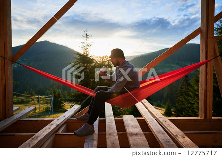 Man on construction site of wooden-framed house sits in hammock against backdrop of mountains and sunrise, sipping coffee. The concept pertains to modern ecological construction. Man on construction site of wooden-framed house sits in hammock against backdrop of mountains and sunrise, sipping coffee. The concept pertains to modern ecological construction. 112775717