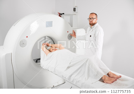 Medical computed tomography or MRI scanner. Male radiologist presses MRI button to examine female patient, looking to the camera. 112775735