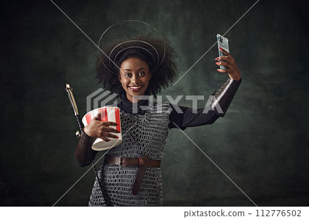 Smiling young African woman, medieval warrior in chainmail armor holding sword and taking selfie with mobile phone against vintage green background. Smiling young African woman, medieval warrior in chainmail armor holding sword and taking selfie with mobile phone against vintage green background. 112776502