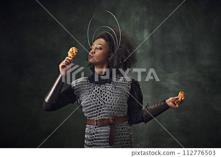 Young African woman, medieval warrior in chainmail armor holding fried chicken against vintage green background. Food ads 112776503