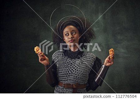 Young African woman, medieval warrior in chainmail armor holding fried chicken against vintage green background. Gourmet food campaign 112776504