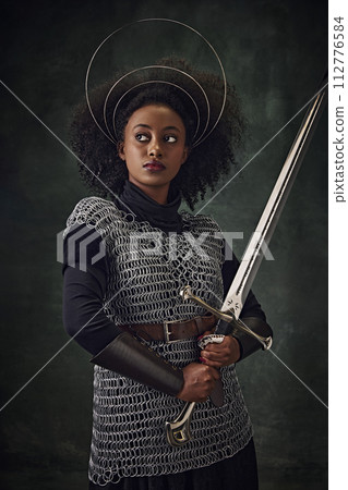 Serious, young African woman, medieval warrior in chainmail armor with halo-like rings above head and sword against vintage green background Serious, young African woman, medieval warrior in chainmail armor with halo-like rings above head and sword against vintage green background 112776584