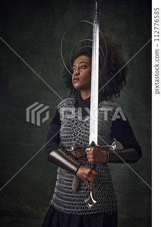Elegant young African-American woman in medieval chainmail armor with halo-like rings above head holding sword against dark green background Elegant young African-American woman in medieval chainmail armor with halo-like rings above head holding sword against dark green background 112776585