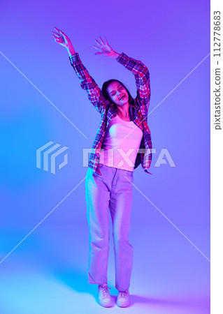 Happy and positive young woman in checkered shirt cheerfully dancing and having fun against purple studio background in neon light 112778683