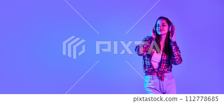 Smiling and relaxed young woman in checkered shirt listening to music in headphones against purple studio background in neon light 112778685