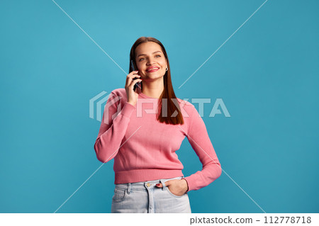 Portrait of beautiful smiling young woman in pink sweater and blue jeans talking on mobile phone against blue studio background Portrait of beautiful smiling young woman in pink sweater and blue jeans talking on mobile phone against blue studio background 112778718
