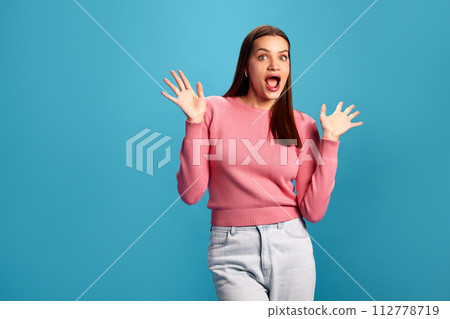 Emotional young woman in pink sweater and jeans expressing positive shock and excitement against blue studio background. Surprise Emotional young woman in pink sweater and jeans expressing positive shock and excitement against blue studio background. Surprise 112778719