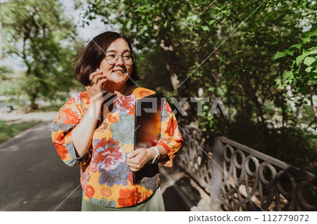 Happy woman chatting on the phone holding a laptop walking in the park Smiling freelancer wearing a colorful blouse using smartphone outdoors Happy woman chatting on the phone holding a laptop walking in the park Smiling freelancer wearing a colorful blouse using smartphone outdoors 112779072