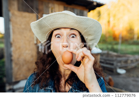 Funny Woman Farmer in denim with a fresh egg, making a funny face. A woman in hat exhibits sheer happiness with a fresh egg in hand, standing on her farm Sustainable lifestyle, organic farmer 112779076