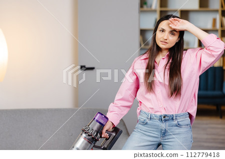 Tired and exhausted woman vacuuming house, cleaning rug carpet in living room, frustrated with doing much work about the house resting for a minute wiping forehead after finishing work, taking break. 112779418
