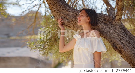 Beautiful Asian young woman in white dress outdoor near olive tree. embracing fresh air and engaging in outdoor activities. Friluftsliv concept means spending as much time outdoors as possible Beautiful Asian young woman in white dress outdoor near olive tree. embracing fresh air and engaging in outdoor activities. Friluftsliv concept means spending as much time outdoors as possible 112779444