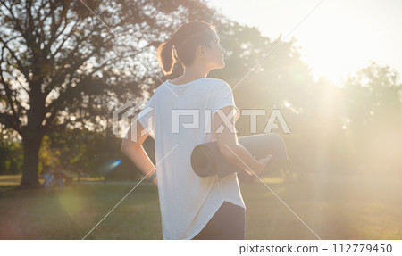 Healthy active lifestyle concept in city park. woman walking with exercise mat through park in summer day, after or before health fitness, wellness exercise or outdoor workout. 112779450