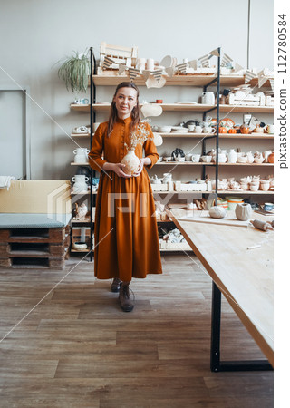 Woman Ceramic Artist Holding Artisan Clay Vase in Sunny Studio. Elegant woman holding a pottery vase with dried flowers in a sunny ceramics workshop 112780584