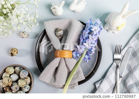 Easter table in gray tones. Plates, napkin, quail eggs, cutlery, flowers, ceramic bunnies. Top view Easter table in gray tones. Plates, napkin, quail eggs, cutlery, flowers, ceramic bunnies. Top view 112780987