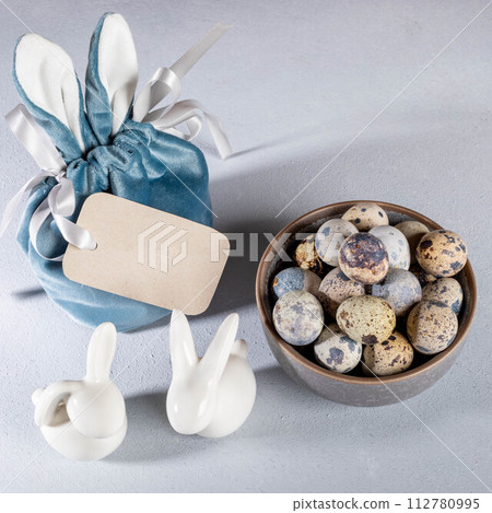 Easter gift bag with bunny ears, blank note, white ceramic bunnies, bowl full of quail eggs on gray. 112780995