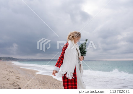 Blond woman holding Christmas tree by the sea. Christmas portrait of a happy woman walking along the beach and holding a Christmas tree in her hands. Dressed in a red coat, white dress. 112782081