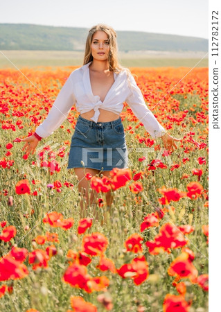 Happy woman in a poppy field in a white shirt and denim skirt with a wreath of poppies on her head posing and enjoying the poppy field. Happy woman in a poppy field in a white shirt and denim skirt with a wreath of poppies on her head posing and enjoying the poppy field. 112782172