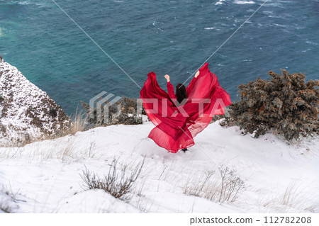 Woman red dress snow sea. Happy woman in a red dress in the snowy mountains by the emerald sea. The wind blows her clothes, posing against sea and snow background. 112782208