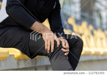 Close-up of a male athlete in sportswear sitting on stadium stands, clutching his knee in discomfort, exemplifying sports injury. Close-up of a male athlete in sportswear sitting on stadium stands, clutching his knee in discomfort, exemplifying sports injury. 112782303