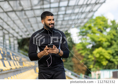 A smiling, bearded man wearing sportswear and earphones holds a smartphone while standing at an outdoor stadium. A smiling, bearded man wearing sportswear and earphones holds a smartphone while standing at an outdoor stadium. 112782332