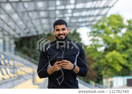 Portrait of a smiling young man taking a break from running, holding a smartphone and wearing earphones in an urban environment. 112782340