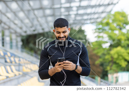 A young, athletic man in sportswear is smiling while using his smartphone and wired earphones at a stadium, conveying a sense of leisure and technology. 112782342