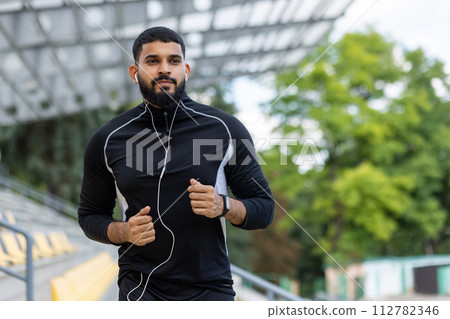 Focused bearded runner with earphones on an urban track. Healthy lifestyle and fitness concept captured in natural daylight. 112782346