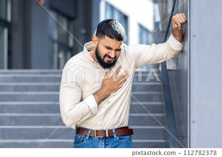 Panic and heart attack on the street. A young Indian man is standing near a building, leaning against a wall and holding his chest with his hand, feeling severe pain and needing medical help. 112782378