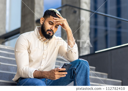 Photo of a sad young Muslim man sitting near a building on the stairs, holding his head and looking worriedly at the mobile phone screen. Photo of a sad young Muslim man sitting near a building on the stairs, holding his head and looking worriedly at the mobile phone screen. 112782410