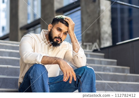 Upset and disappointed young Indian man sitting on the steps of an office building and holding his hand above his head, looking thoughtfully to the side. Upset and disappointed young Indian man sitting on the steps of an office building and holding his hand above his head, looking thoughtfully to the side. 112782412