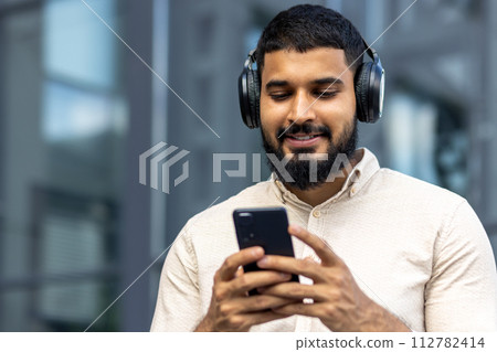A cheerful young man with a beard is seen outside using his smartphone and listening to music through over-ear headphones, exuding a vibe of relaxation and enjoyment in an urban setting. A cheerful young man with a beard is seen outside using his smartphone and listening to music through over-ear headphones, exuding a vibe of relaxation and enjoyment in an urban setting. 112782414