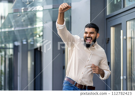 Indian young man standing near an office building and rejoicing in success raising his hand up and shouting with happiness, he got a job, a promotion, a business deal. 112782423