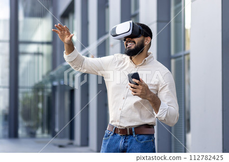 Photo of a young Muslim man in a shirt standing outside an office building wearing a virtual mask, holding a phone, talking online and gesturing with his hands. Photo of a young Muslim man in a shirt standing outside an office building wearing a virtual mask, holding a phone, talking online and gesturing with his hands. 112782425