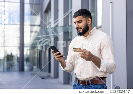 Confident young adult male using a smartphone and holding a credit card on a sunny day outside modern office buildings. Concept of mobile banking, e-commerce and modern lifestyle. Confident young adult male using a smartphone and holding a credit card on a sunny day outside modern office buildings. Concept of mobile banking, e-commerce and modern lifestyle. 112782447