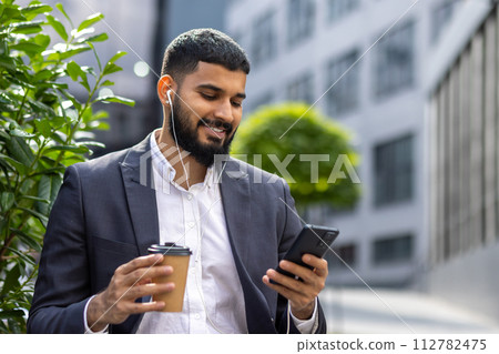 A cheerful young businessman wearing a suit is enjoying music with headphones and holding a coffee cup while using a smartphone ,outside an office building surrounded by greenery. A cheerful young businessman wearing a suit is enjoying music with headphones and holding a coffee cup while using a smartphone ,outside an office building surrounded by greenery. 112782475