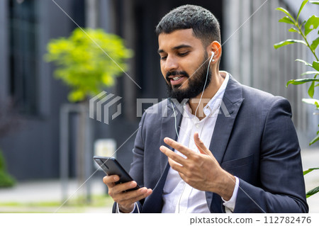 A well-dressed man engages with his smartphone while using earphones outside a modern building, possibly during a work break or in transit. A well-dressed man engages with his smartphone while using earphones outside a modern building, possibly during a work break or in transit. 112782476