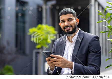 Close-up portrait of a smiling young Muslim businessman in a suit standing outside an office building wearing headphones, holding a phone and looking confidently at the camera. 112782481