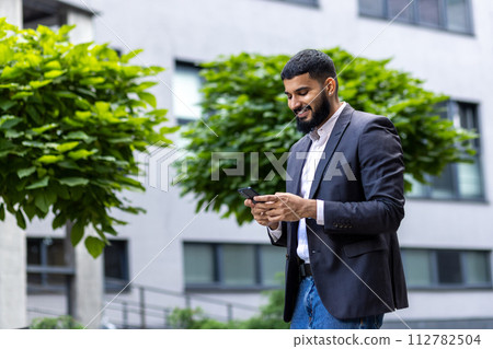 A young, smartly dressed businessman engages with his smartphone while standing outdoors. The image encapsulates themes of connectivity, entrepreneurship, and modern communication. 112782504