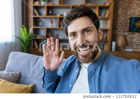 Happy adult male smiling and waving at the camera, likely engaging in a casual video call from a cozy living room setting. 112782612