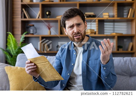 A perplexed adult male sits on a sofa at home, holding an envelope and a letter with a look of confusion and concern. A perplexed adult male sits on a sofa at home, holding an envelope and a letter with a look of confusion and concern. 112782621